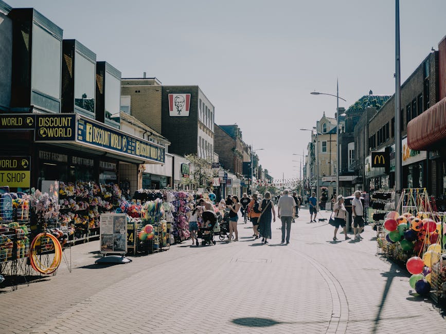 A lively urban street scene with pedestrians walking along a paved shopping promenade lined with various retail stores and snack shops, including a prominent KFC outlet on the left. The street feature